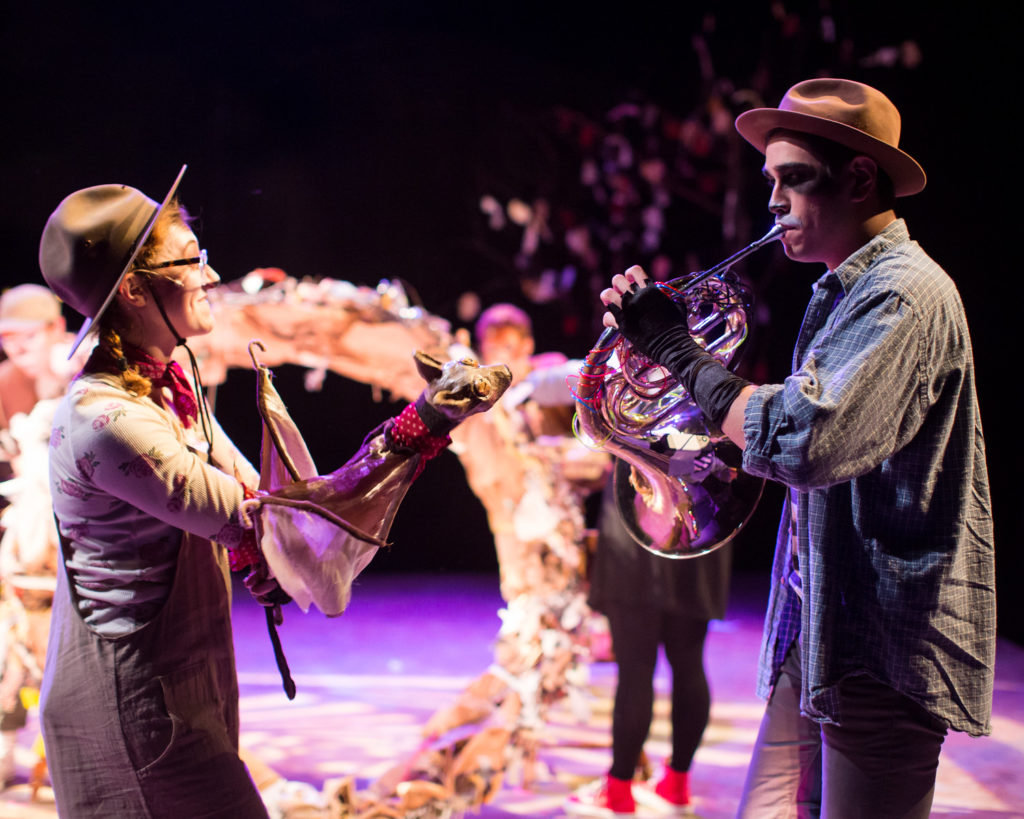 Production photo from ImagineU's production of "Stellaluna." In photo's foreground: Stellaluna puppet operated by female performer while male performer plays french horn. In background is bird's nest. Thrust stage is washed in purple with white light putting focus on performers.